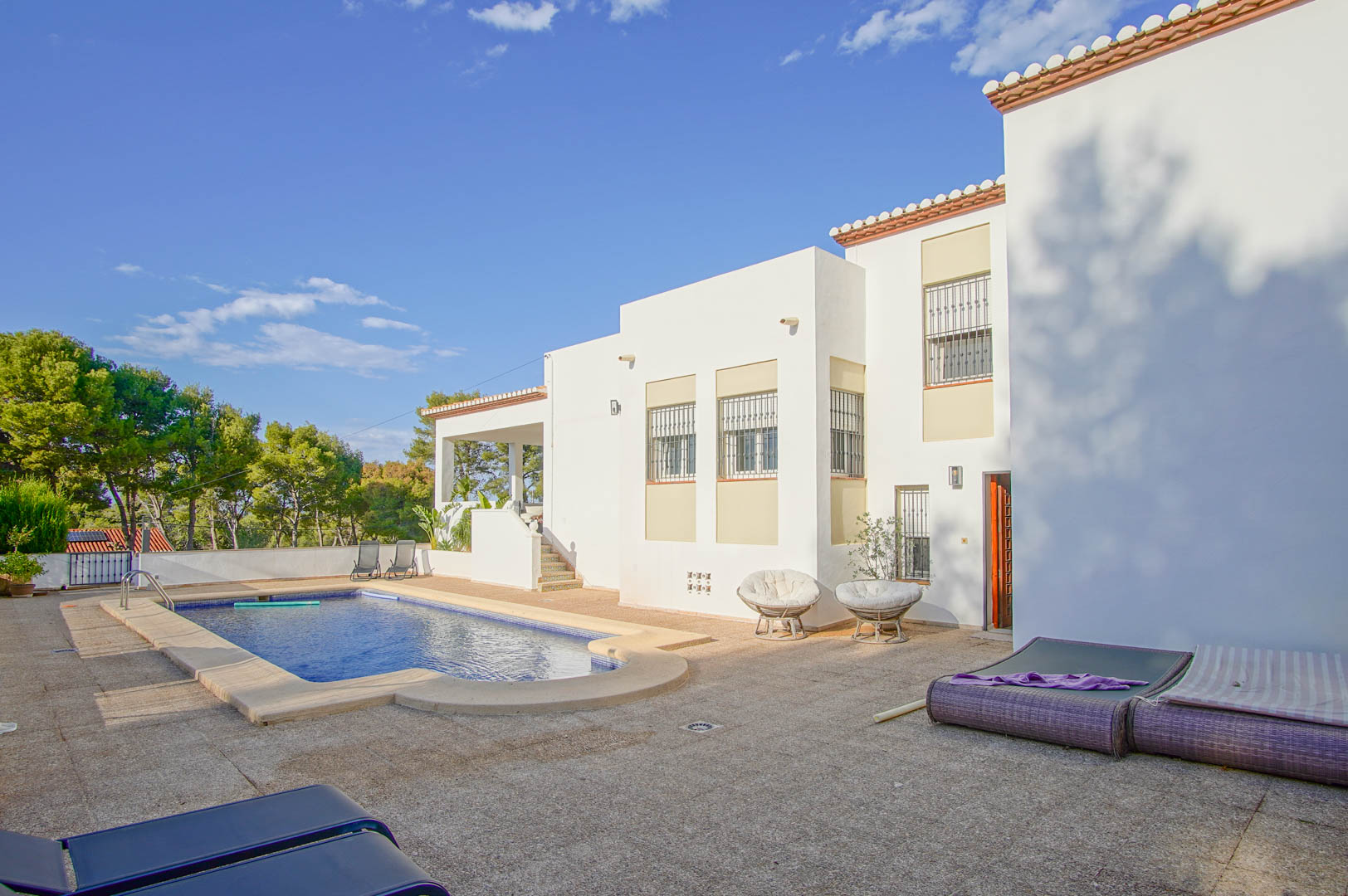 Buitenaanzicht van de witte villa met privézwembad en een ruim betegeld terras, omgeven door mediterrane vegetatie en pijnbomen.