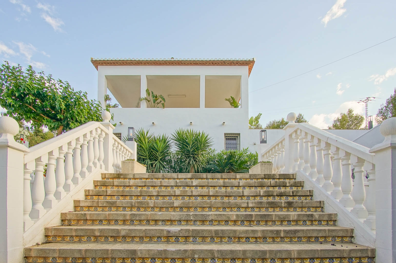 Buitentrap van natuursteen met decoratieve keramische tegels en een witte balustrade, leidend naar het overdekte terras en de gevel van de villa.