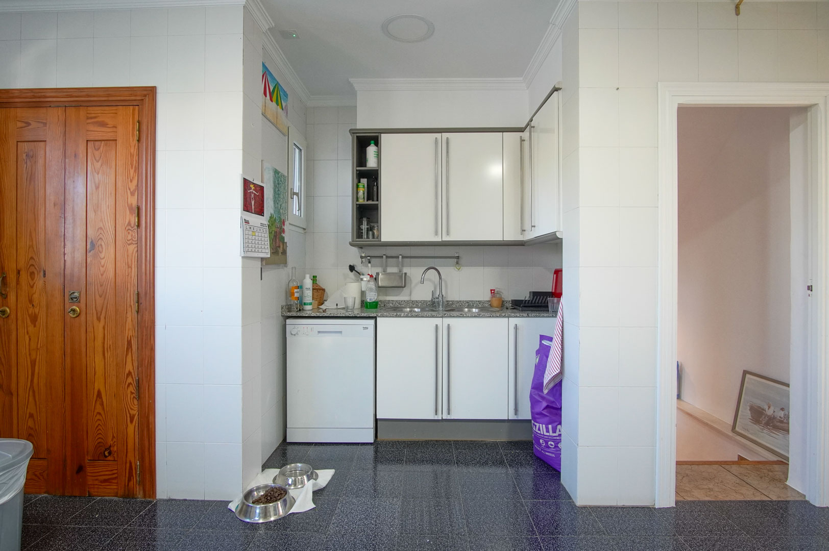 Functionele keuken van het gastenverblijf met witte kasten, een granieten werkblad en donkere terrazzovloer, gelegen naast de toegang tot de bovenverdiepingen.
