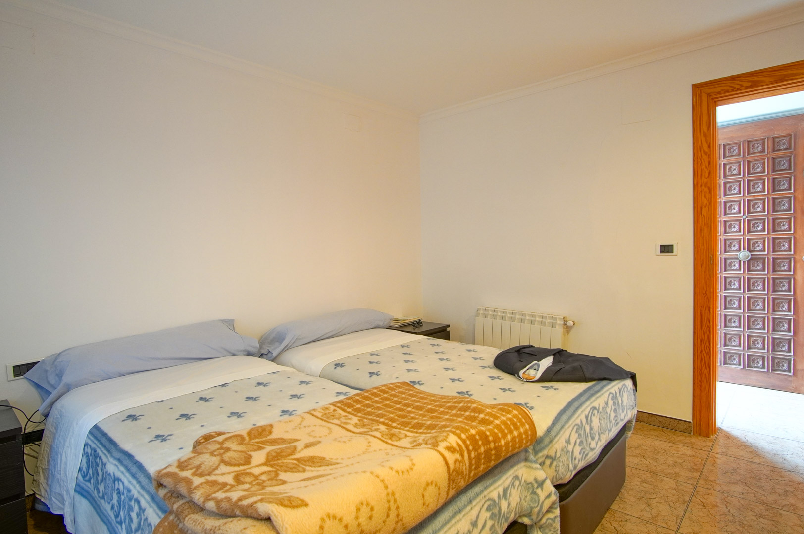 Functionele slaapkamer met twee bedden, witte muren en een tegelvloer. Voorzien van een radiator en een opvallende houten deur met houtsnijwerk.