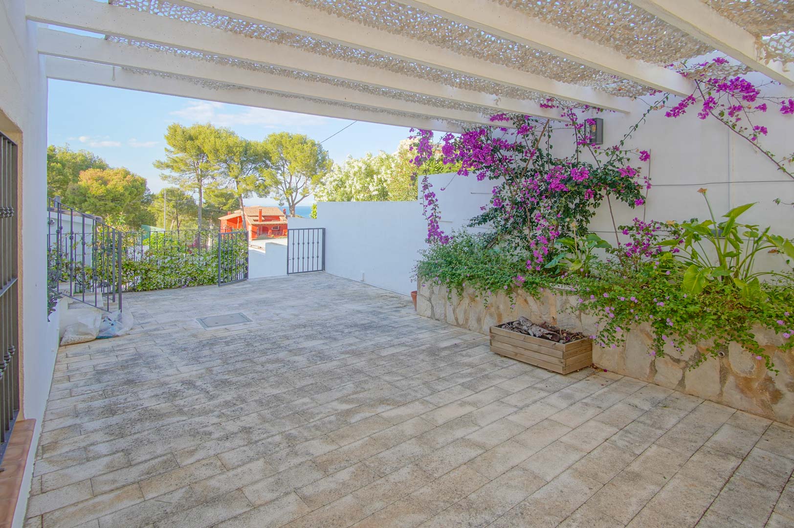 Geplaveid buitenterras met een witte pergola en schaduwnet, voorzien van stenen plantenbakken met bougainville en uitzicht op de omringende natuur en de zee.