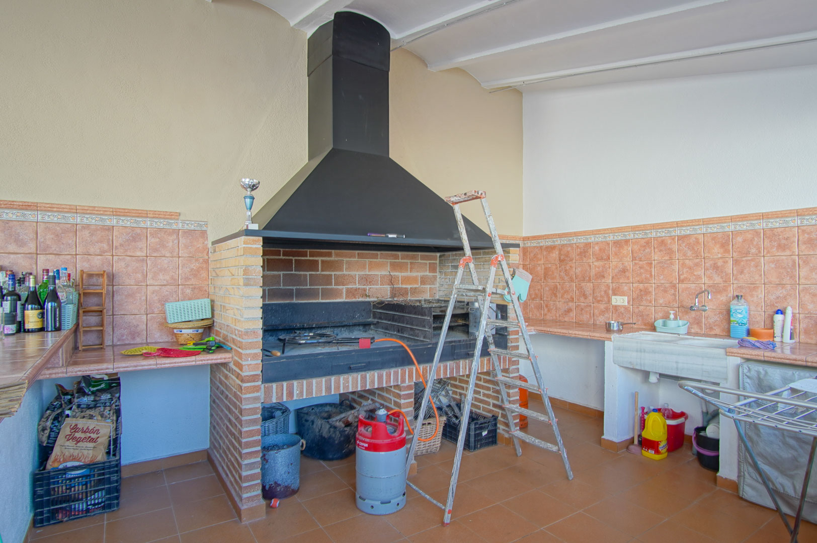 Ingebouwde bakstenen barbecueplaats met een grote zwarte metalen kap, stenen gootsteen en betegelde werkbladen in terracottakleuren.