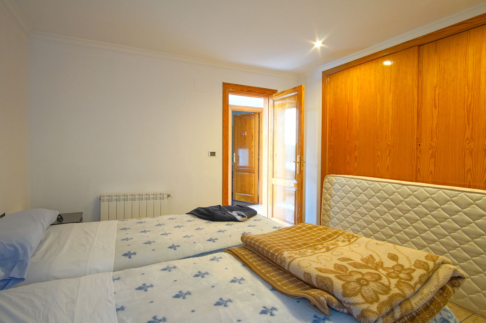 Lichte slaapkamer met grote houten inbouwkasten, twee eenpersoonsbedden en een wandradiator. De bijpassende houten deuren en inbouwspots creëren een warme sfeer.