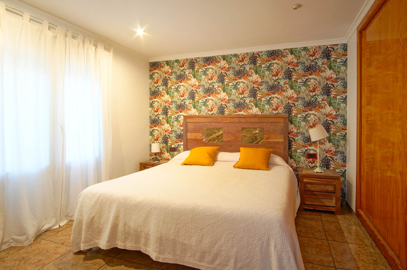 Lichte slaapkamer met tropisch behang, een houten hoofdbord met stenen inlays en ingebouwde houten kasten.