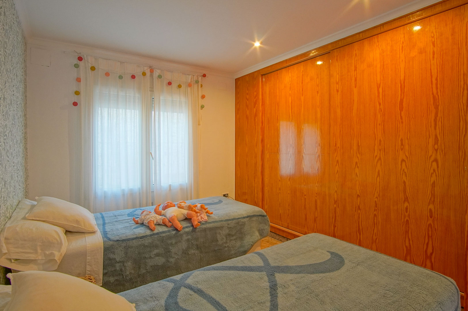 Lichte slaapkamer met twee eenpersoonsbedden, bloemenbehang en een grote ingebouwde houten kledingkast met glanzende afwerking.