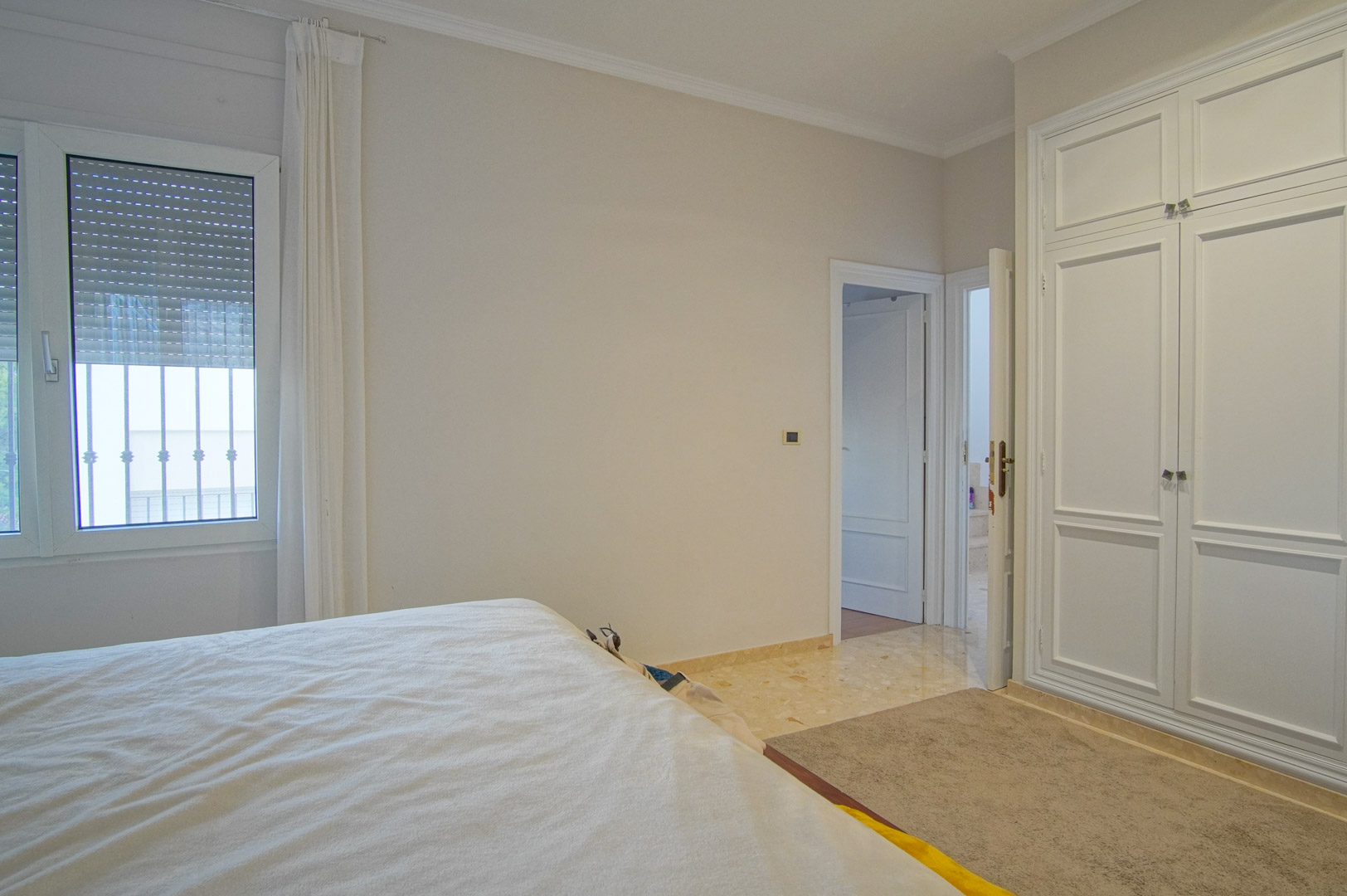 Lichte slaapkamer met witte inbouwkasten, marmeren vloer en raam met rolluiken. Het neutrale kleurenpalet zorgt voor een rustige en elegante uitstraling.