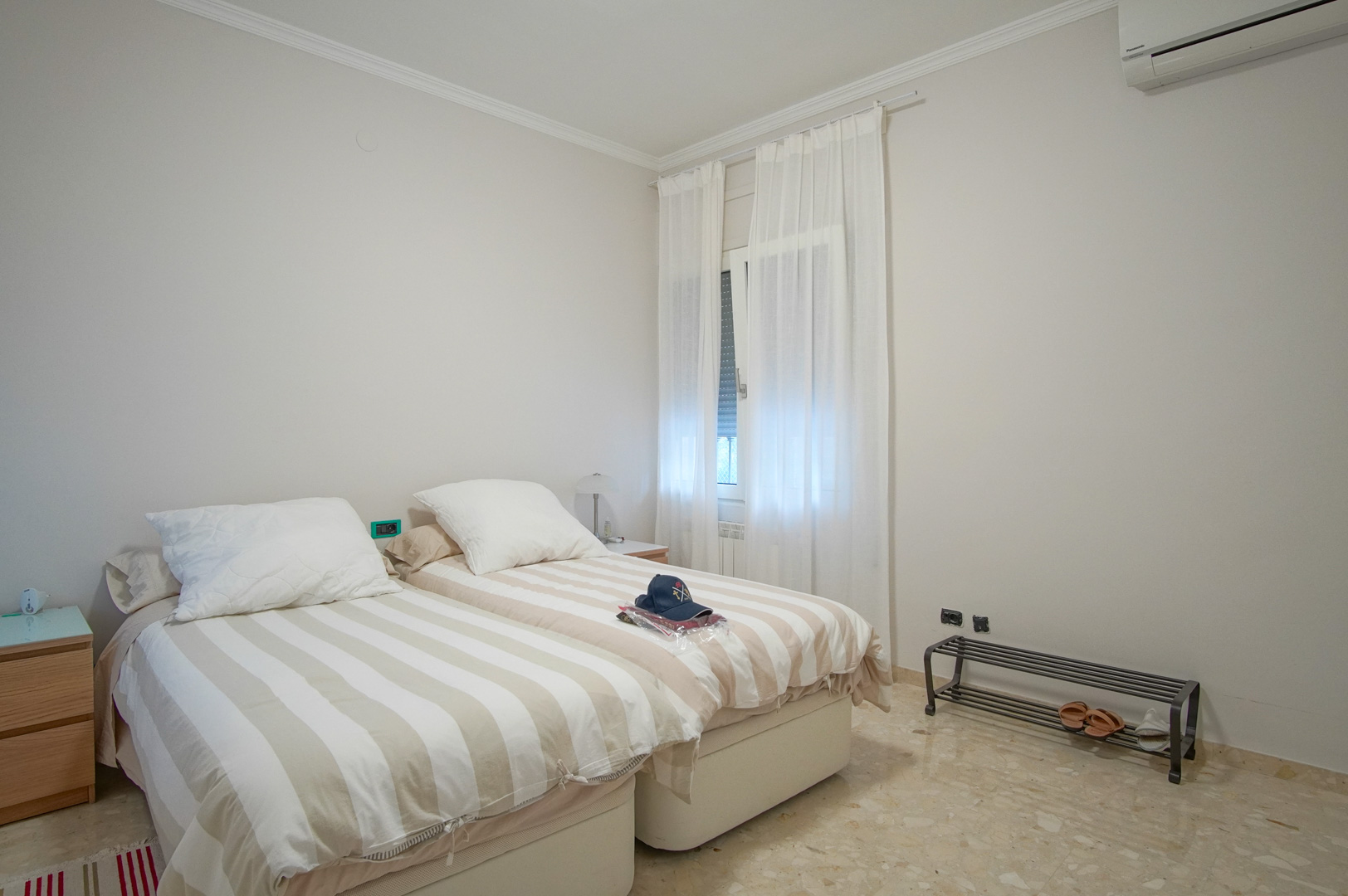Lichte slaapkamer voorzien van twee eenpersoonsbedden, marmeren vloer, airconditioning en een raam met witte gordijnen voor natuurlijk licht.