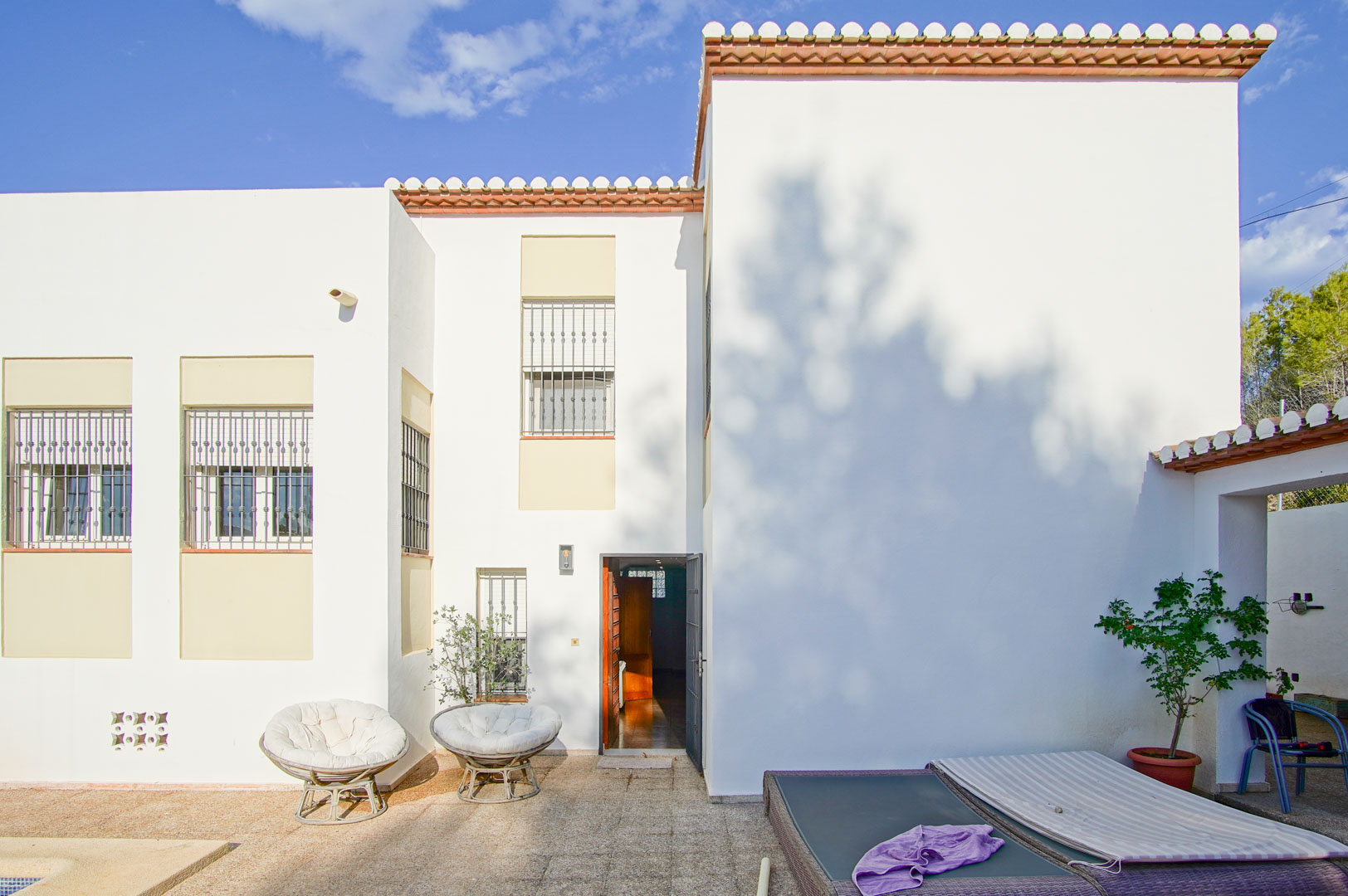 Mediterrane witte gevel met tralies voor de ramen, een stenen terras met buitenmeubilair en directe toegang tot het interieur van de woning.