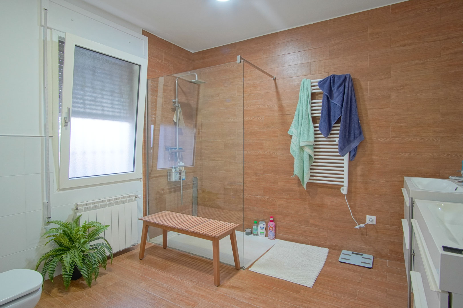 Moderne badkamer met houtlook tegels, een inloopdouche met glazen wand, dubbele wastafel en een witte handdoekradiator.