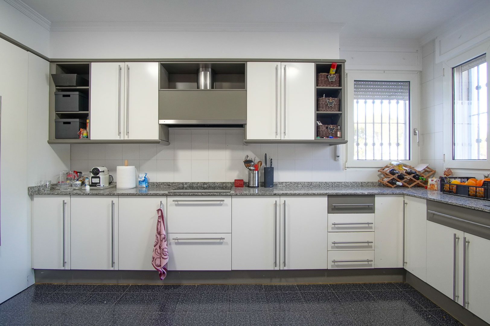 Onafhankelijke keuken met witte kasten, grijs granieten werkblad en inductiekookplaat. Voorzien van een donkere gespikkelde tegelvloer en veel natuurlijk licht.
