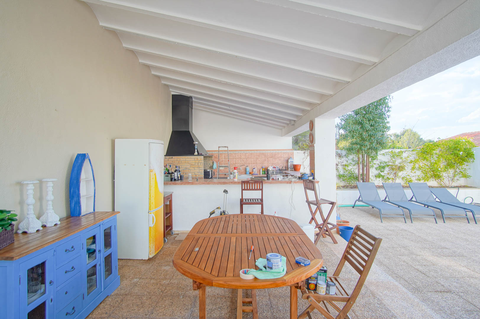 Overdekt terras met buitenkeuken, ingebouwde barbecue en wit balkenplafond. Inclusief houten eethoek met zicht op de ligstoelen bij het zwembad en de tuin.