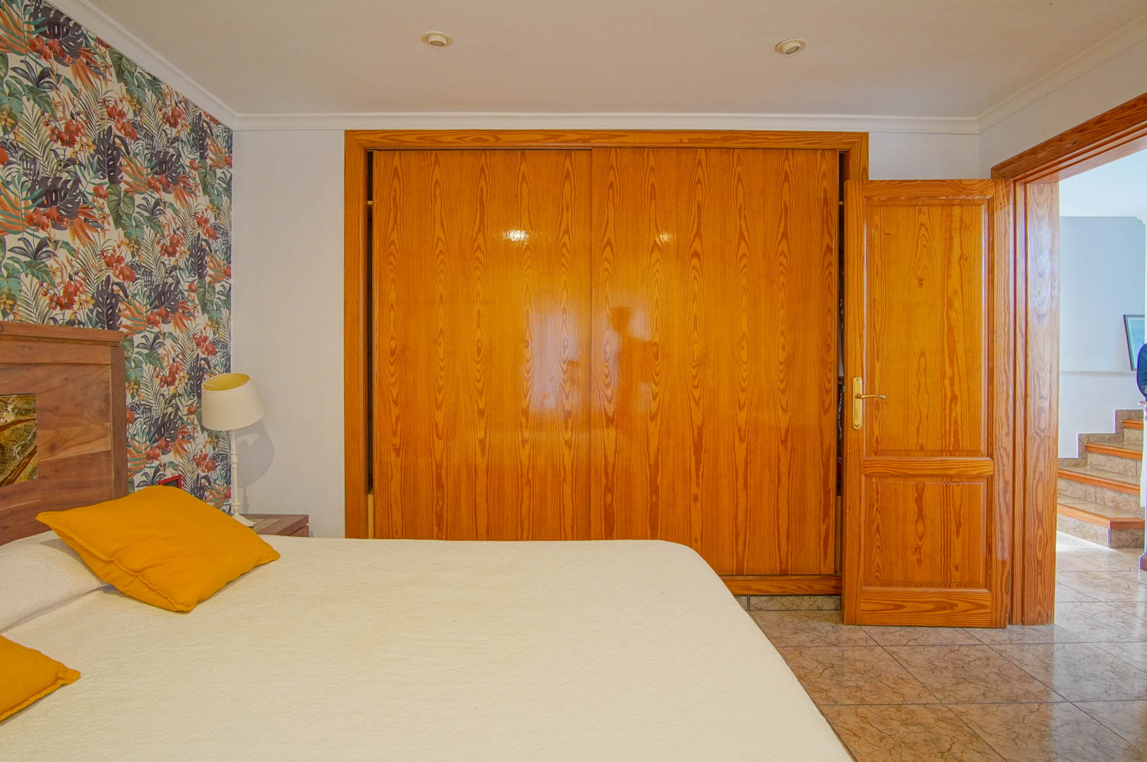 Slaapkamer met een grote houten inbouwkast, een accentmuur met tropisch behang en een tegelvloer. De open deur leidt naar de gang met trap.