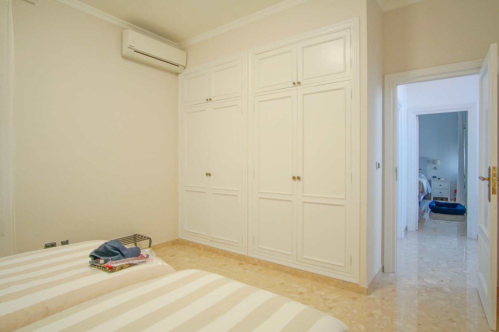 Slaapkamer met grote witte inbouwkasten, marmeren vloer en wandgemonteerde airconditioning, met doorgang naar de centrale gang.