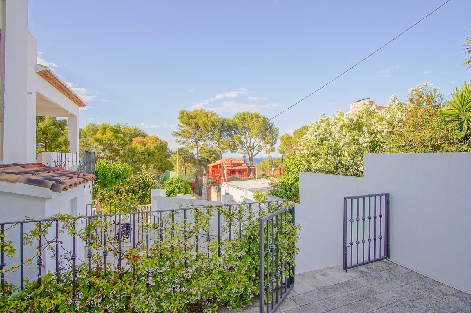 Uitzicht vanaf het stenen terras met smeedijzeren hekwerk en klimplanten, uitkijkend over de mediterrane tuin en de zee in de verte tussen de pijnbomen van Dénia.