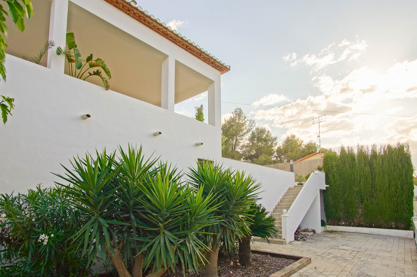 Witte mediterrane gevel met overdekt terras en vierkante zuilen. De buitenruimte is voorzien van een betegelde trap en een tuin met yucca's en een cipressenhaag.