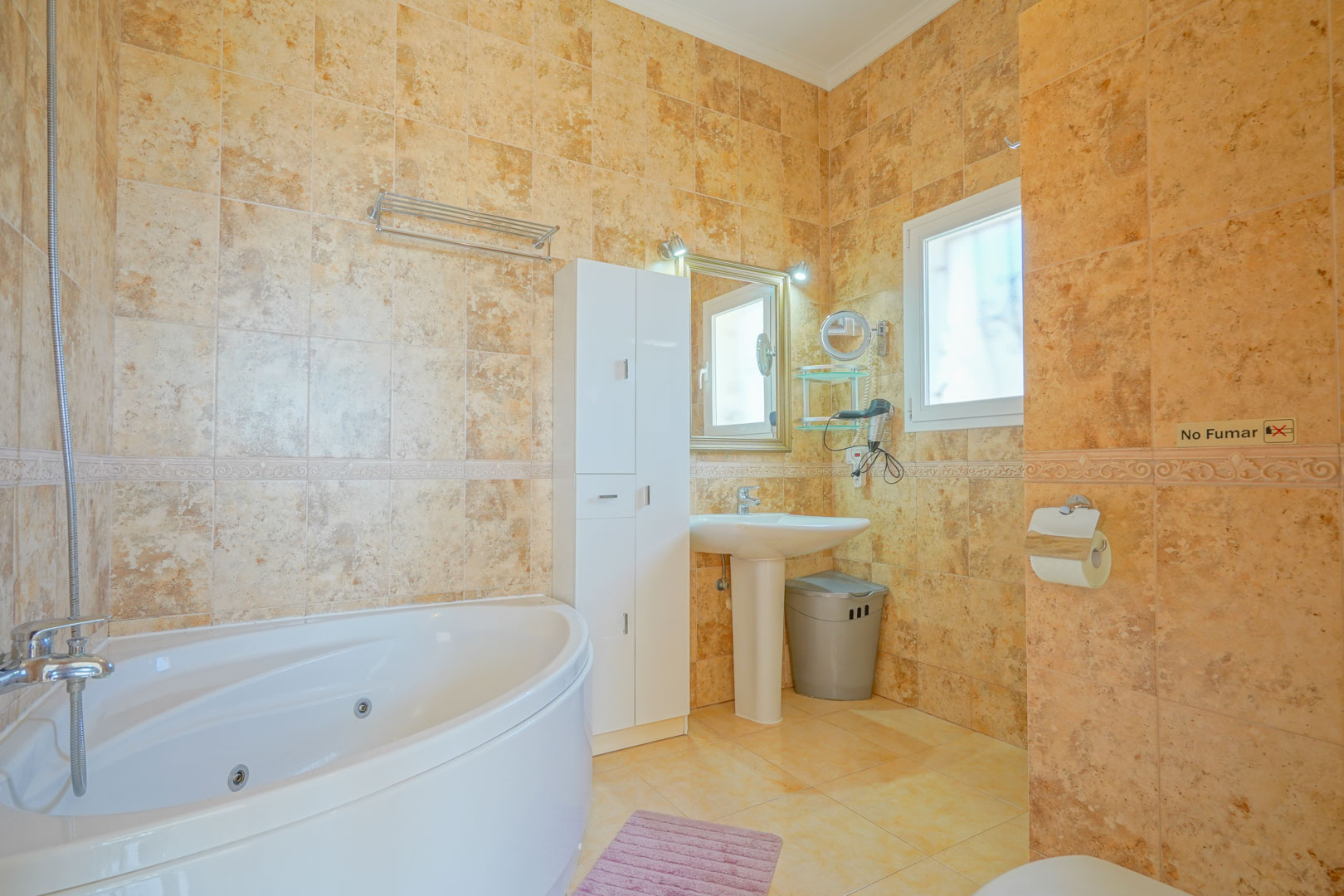 Badkamer met een hoek-whirlpoolbad, warmgekleurde keramische wandtegels, een wastafel op zuil en een witte opbergkast.