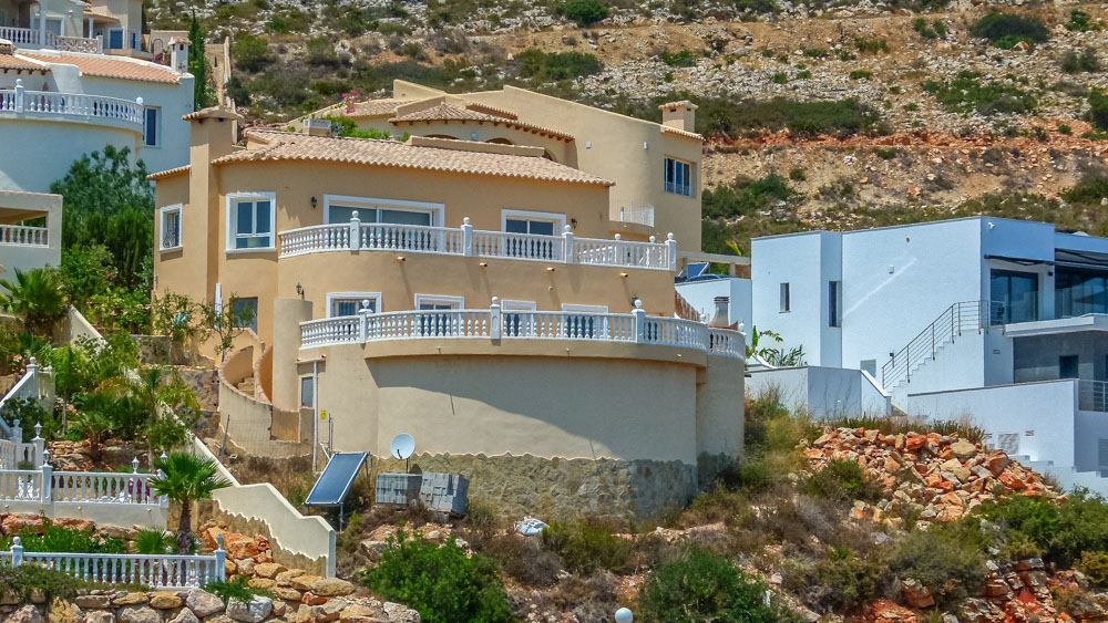 Exterieur van de mediterrane villa met okerkleurige gevels en royale gebogen terrassen met witte balustrades, gelegen tegen de rotsachtige helling.