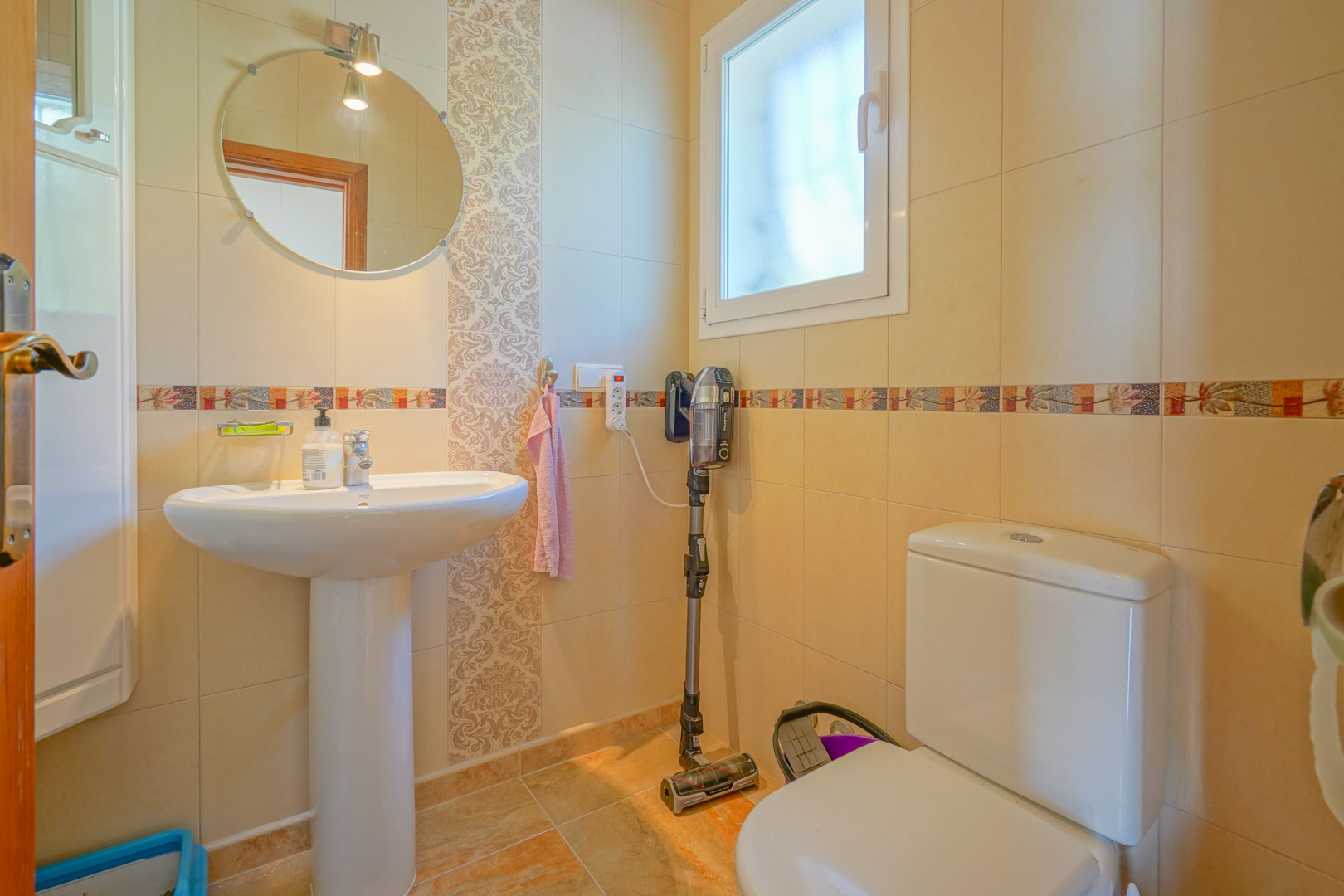 Functionele badkamer met een kolomwastafel, ronde spiegel en crèmekleurige tegelwanden voorzien van decoratieve sierstrips.