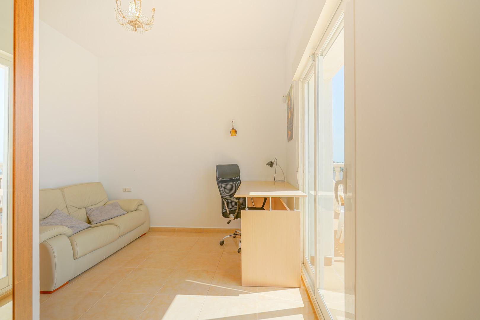 Lichte extra kamer met werkplek, lederen bank en directe toegang tot het terras via grote schuifdeuren. De warme tegelvloer wordt verlicht door overvloedig natuurlijk daglicht.