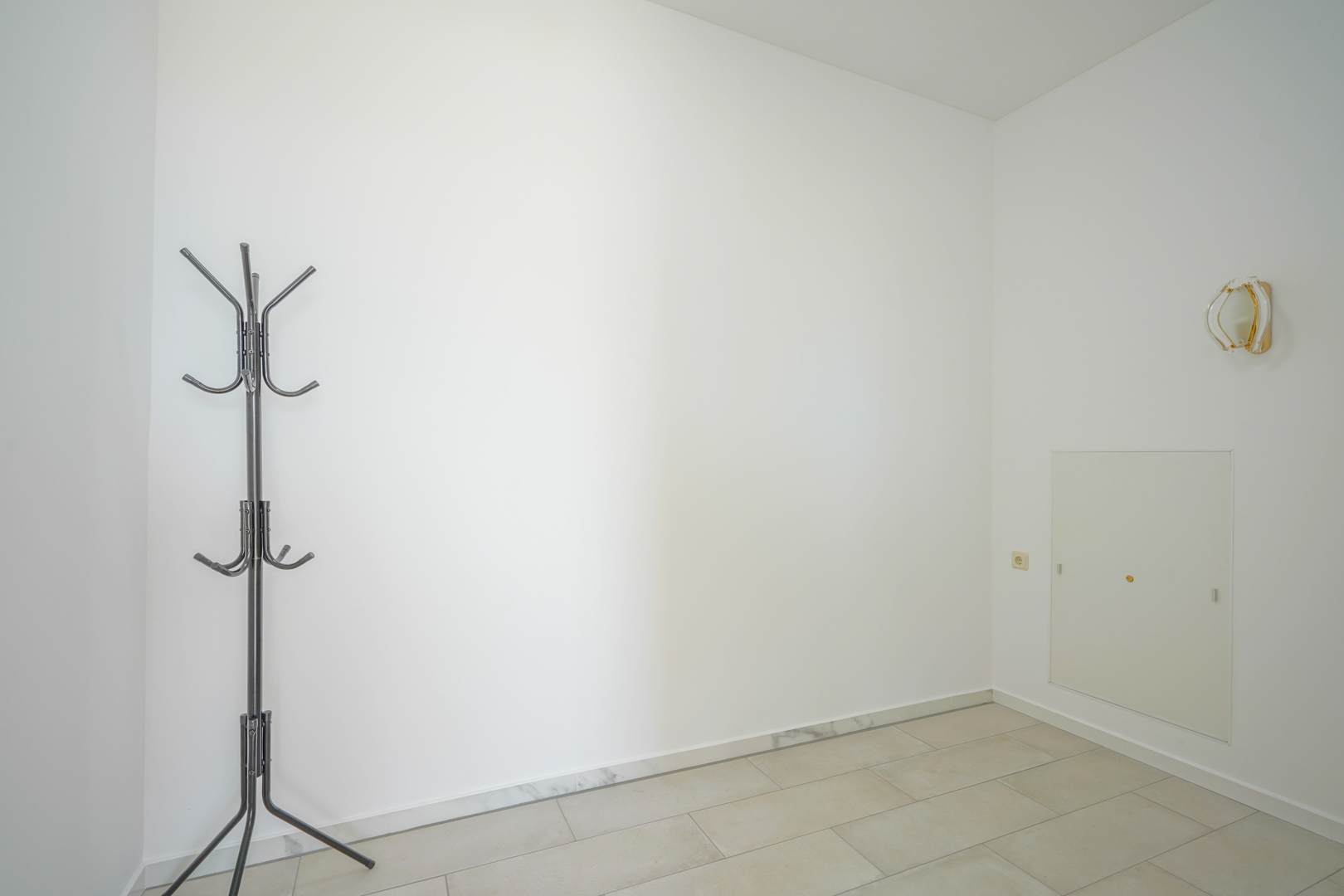 Minimalistische entree met witte muren, lichte tegelvloer met marmeren plinten, een metalen kapstok en een decoratieve wandlamp.
