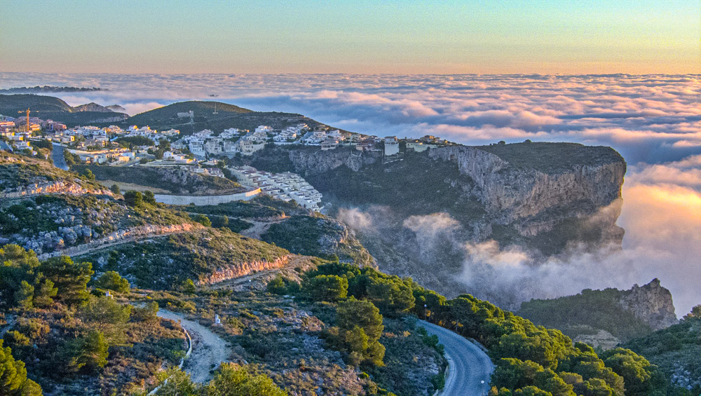 Panoramisch uitzicht op de kliffen van Cumbre del Sol boven een wolkenzee bij zonsondergang, met de ruige kustlijn en de residentiële bebouwing.
