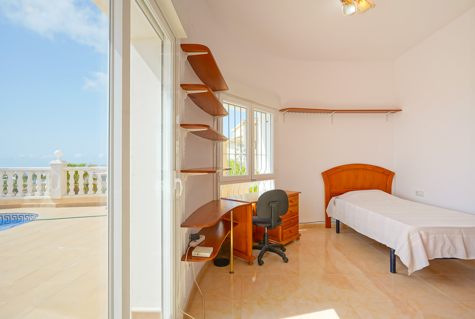 Slaapkamer met eenpersoonsbed, houten bureau en wandplanken. Grote schuifdeuren bieden direct toegang tot het terras bij het zwembad.