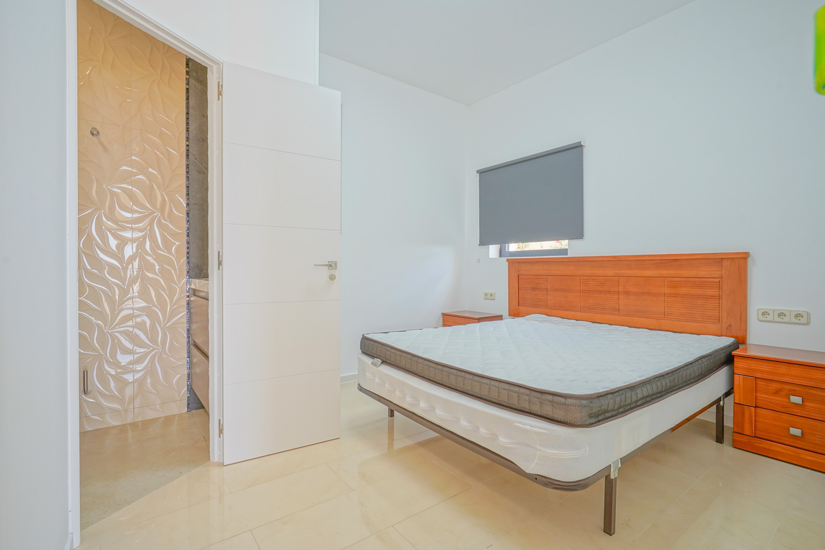 Tweepersoonsslaapkamer met houten meubilair, lichte tegelvloer en een en-suite badkamer met opvallende wandtegels met reliëfpatroon.
