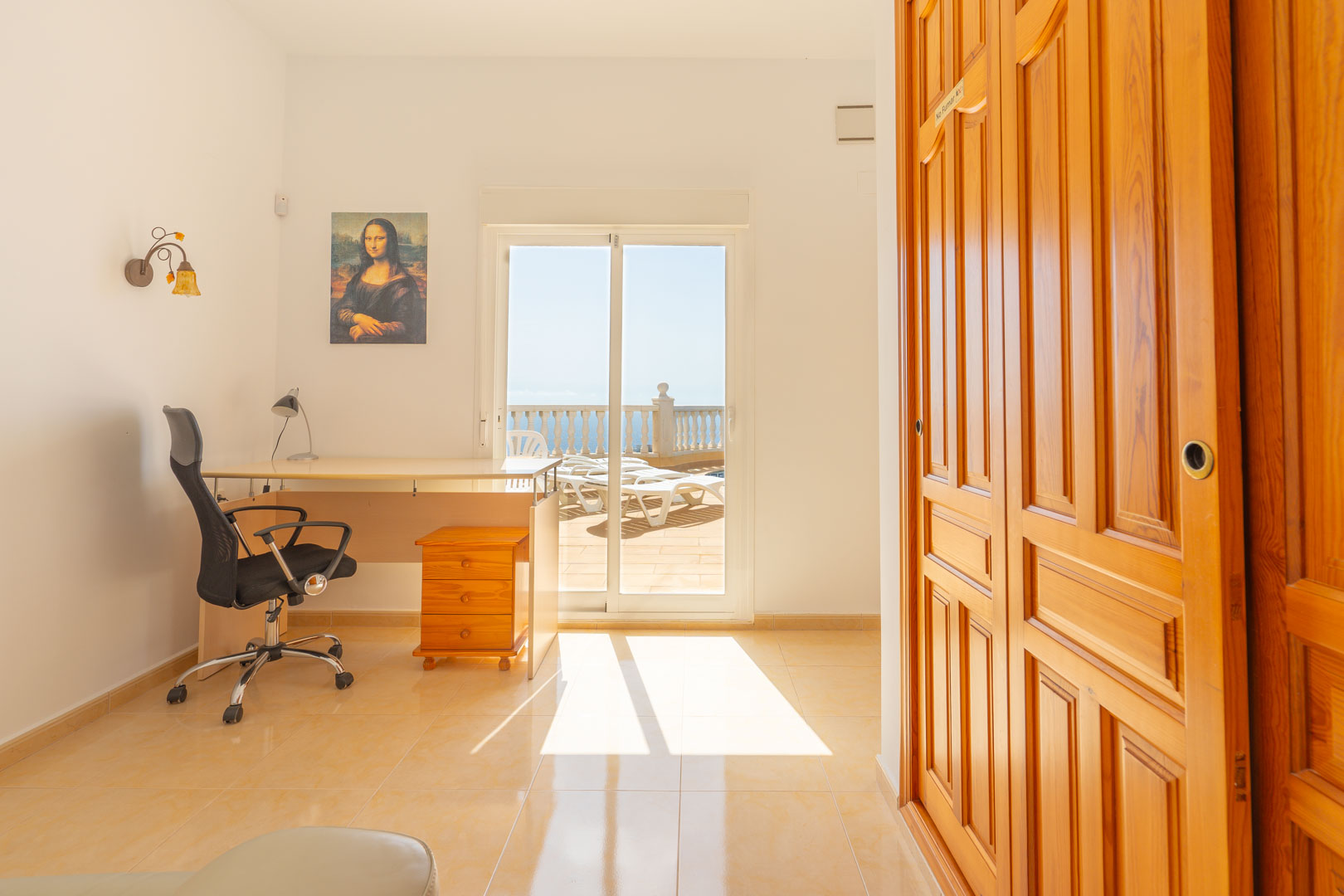 Veelzijdige kamer met werkplek, houten inbouwkasten en directe toegang tot het zonnige terras met uitzicht op zee via een glazen schuifdeur.