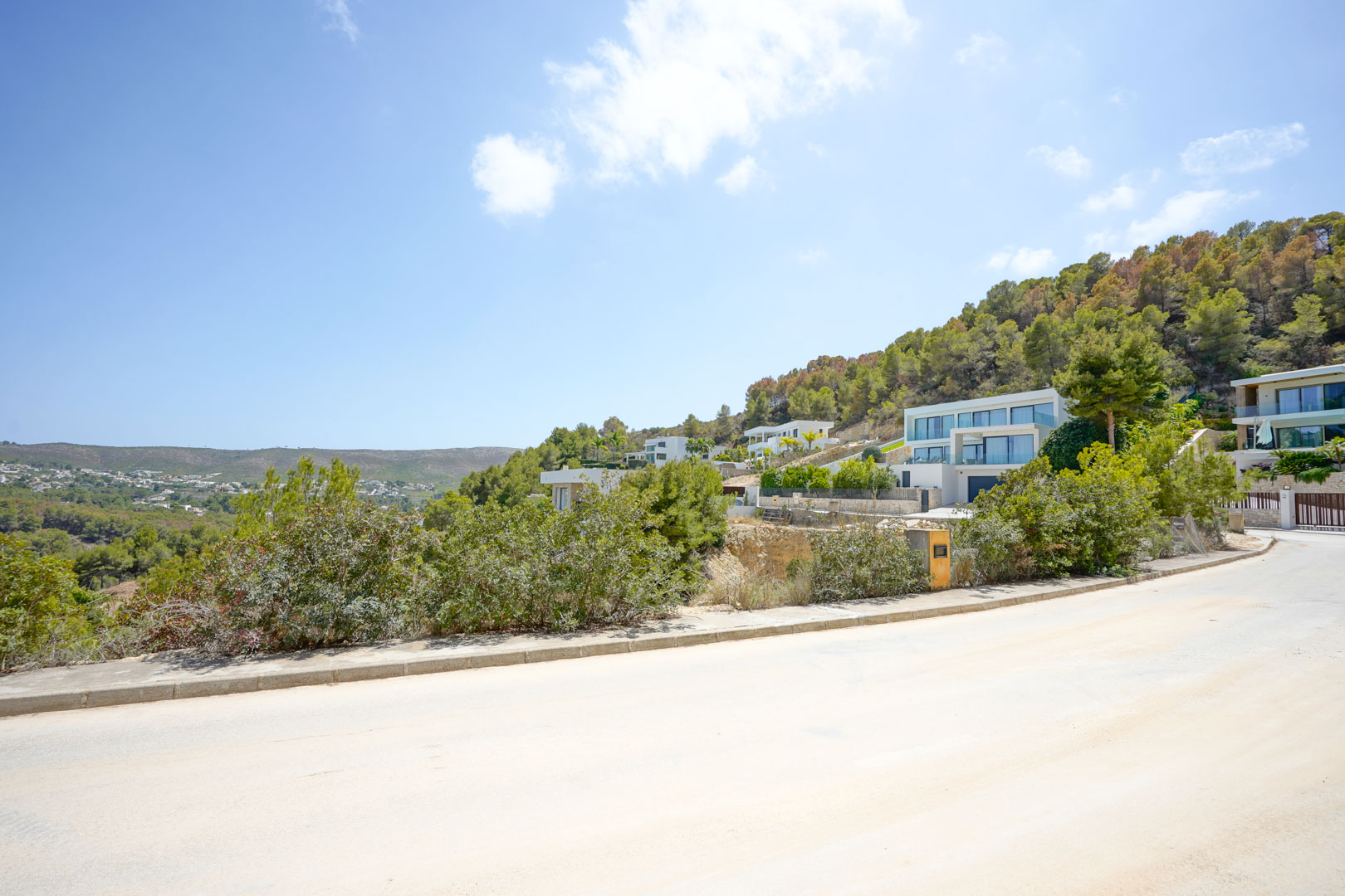 Geasfalteerde toegangsweg in een moderne woonwijk met hedendaagse villa's op een helling, omgeven door mediterraan dennenbos en weidse vergezichten.
