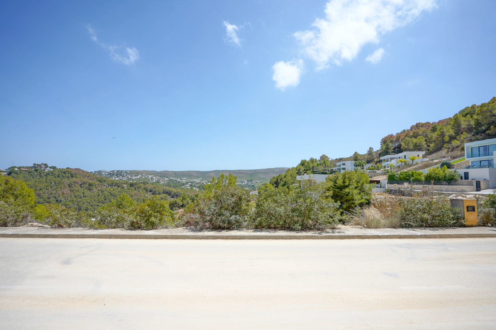Panoramisch uitzicht vanaf de geasfalteerde toegangsweg over de beboste heuvels en moderne villa's, met de volledige infrastructuur en natuurlijke omgeving.