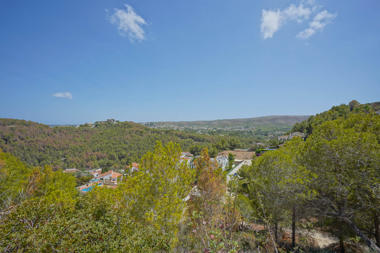 Verhoogd panoramisch uitzicht over een groene vallei met mediterrane pijnbomen, witte villa's met zwembaden en glooiende heuvels onder een helderblauwe lucht.