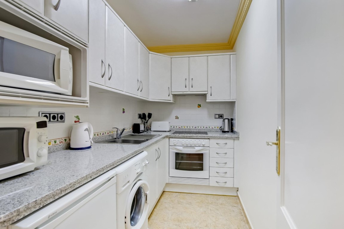 Functionele keuken met witte kasten, grijs granieten werkblad en inbouwapparatuur. Inclusief wasmachine en decoratieve gele plafondlijsten.