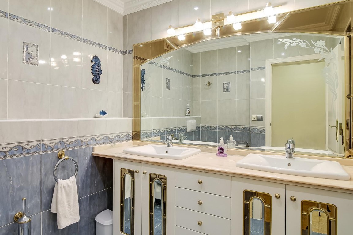 Hoofdbadkamer met dubbele wastafel op een marmeren blad, kasten met gouden details, grote spiegel en decoratieve blauw-witte wandtegels.