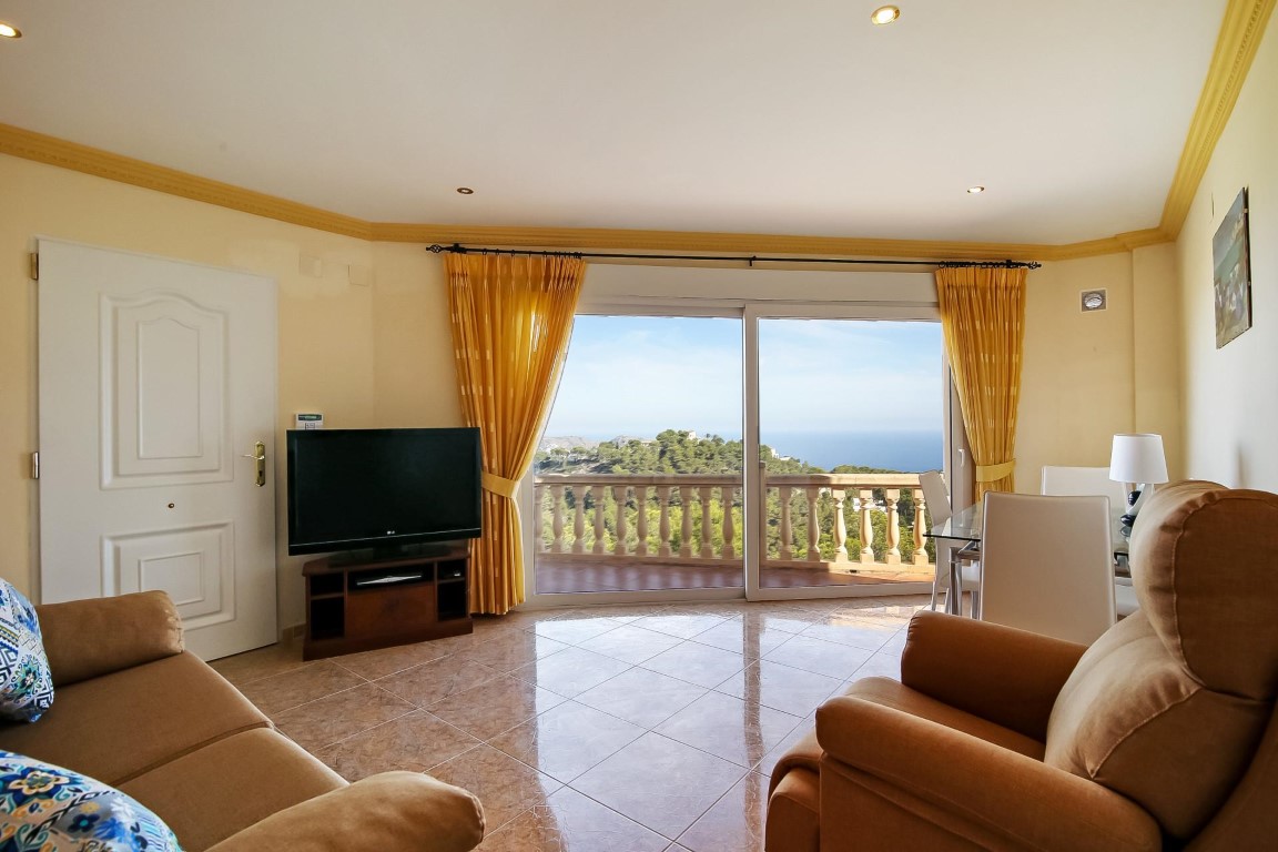 Lichte woonkamer met tegelvloer, sierlijsten en grote schuifpui die een panoramisch uitzicht biedt over de Middellandse Zee en de omliggende heuvels.