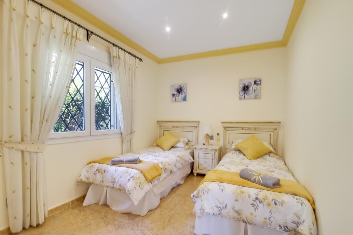 Slaapkamer met twee eenpersoonsbedden, witte klassieke hoofdborden, inbouwspots en een raam voorzien van veiligheidstralies.