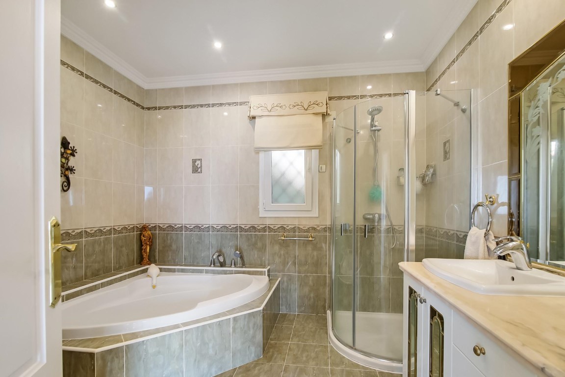 Volledige badkamer met hoekbad, halfronde douchecabine en wastafelmeubel met marmeren blad. Betegelde wanden met sierranden en inbouwspots in het plafond.