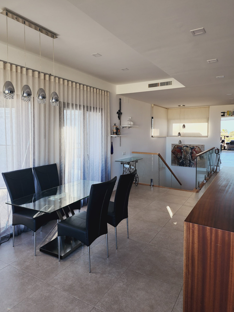 Eethoek met een glazen tafel en zwarte stoelen, grenzend aan een trap met glazen balustrade. Moderne hanglampen en inbouwspots verlichten de ruimte met tegelvloer.