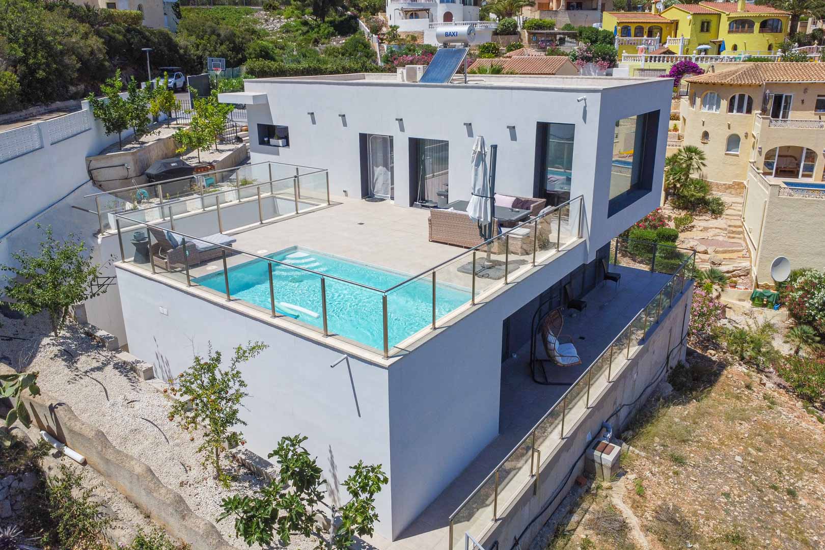 Exterieur van de moderne villa met zwembad op het boventerras, glazen balustrades, zonnepanelen op het dak en een overdekt terras op de benedenverdieping.