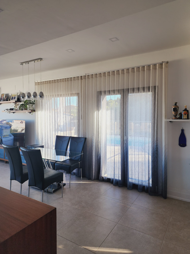 Lichte eethoek met een glazen tafel en hoge rugstoelen, gelegen bij grote ramen met kleurverloopgordijnen die directe toegang bieden tot het terras bij het zwembad.