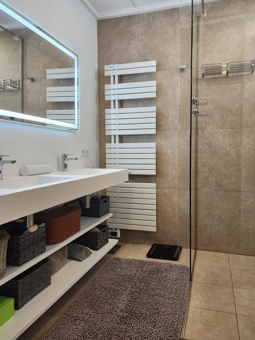 Moderne badkamer met dubbele wastafel, open schappen en een LED-verlichte spiegel. Voorzien van een witte handdoekradiator en een inloopdouche met aardetinten tegels.