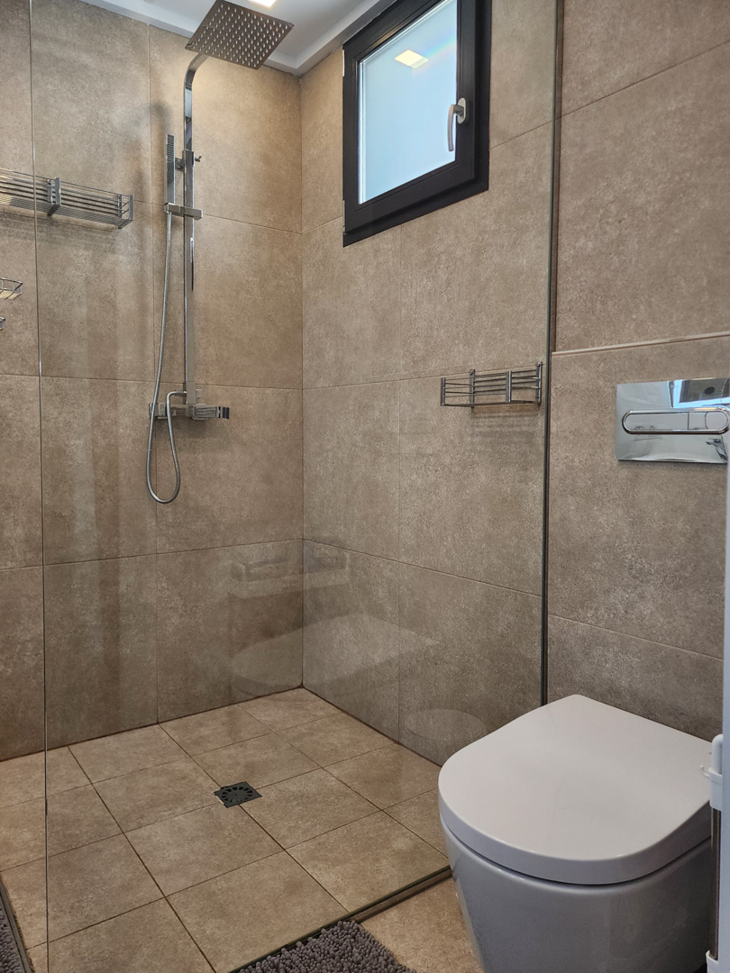 Moderne badkamer met inloopdouche, glazen wand, chromen regendouche en keramische tegels in natuurlijke zandkleuren.