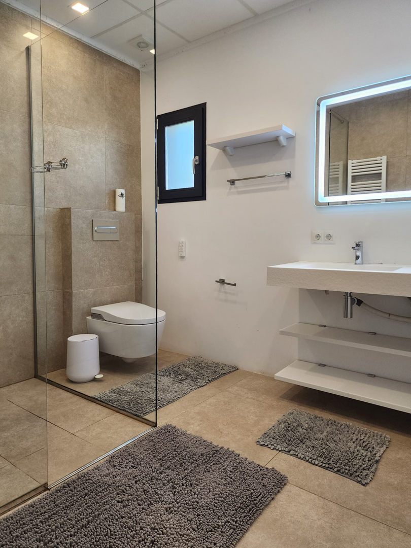 Moderne badkamer met inloopdouche, glazen wand, hangend toilet en zwevende wastafel met open schappen en een spiegel met LED-achtergrondverlichting.