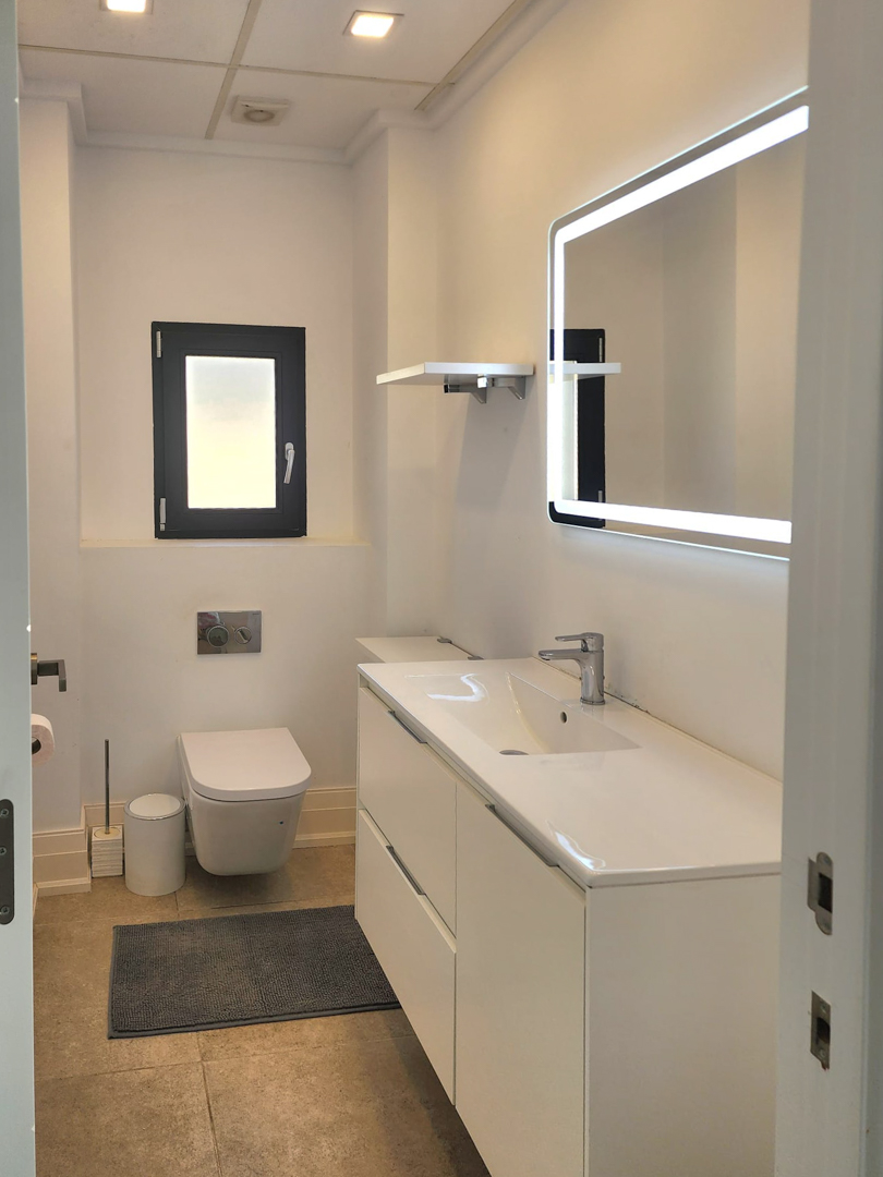 Moderne badkamer met wit wastafelmeubel, LED-verlichte spiegel en hangend toilet. Voorzien van inbouwspots in het plafond en neutrale vloertegels.