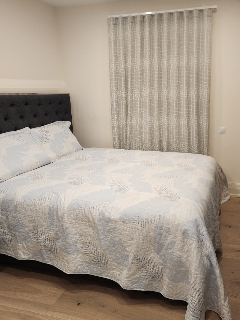 Moderne slaapkamer met tweepersoonsbed, donkergrijs gecapitonneerd hoofdbord, lichte houten vloer en kamerhoge gordijnen met patroon.
