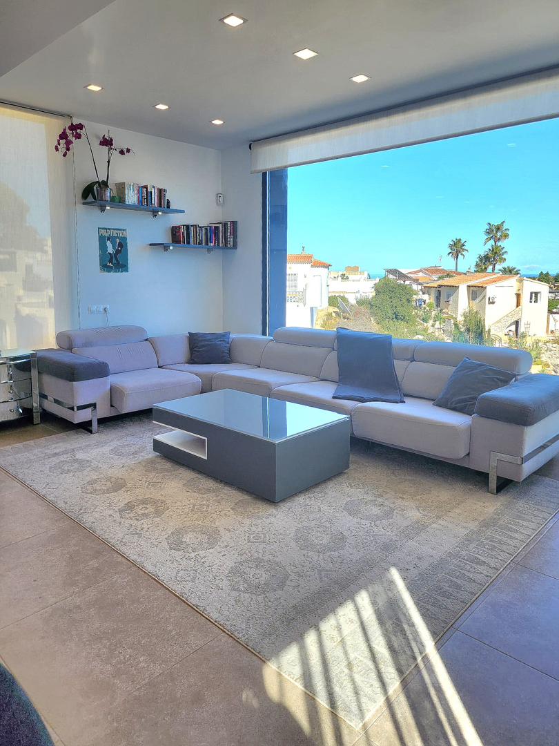 Moderne woonkamer met L-vormige bank, minimalistische salontafel en groot panoramaraam. Voorzien van inbouwspots, zwevende planken en een tegelvloer.