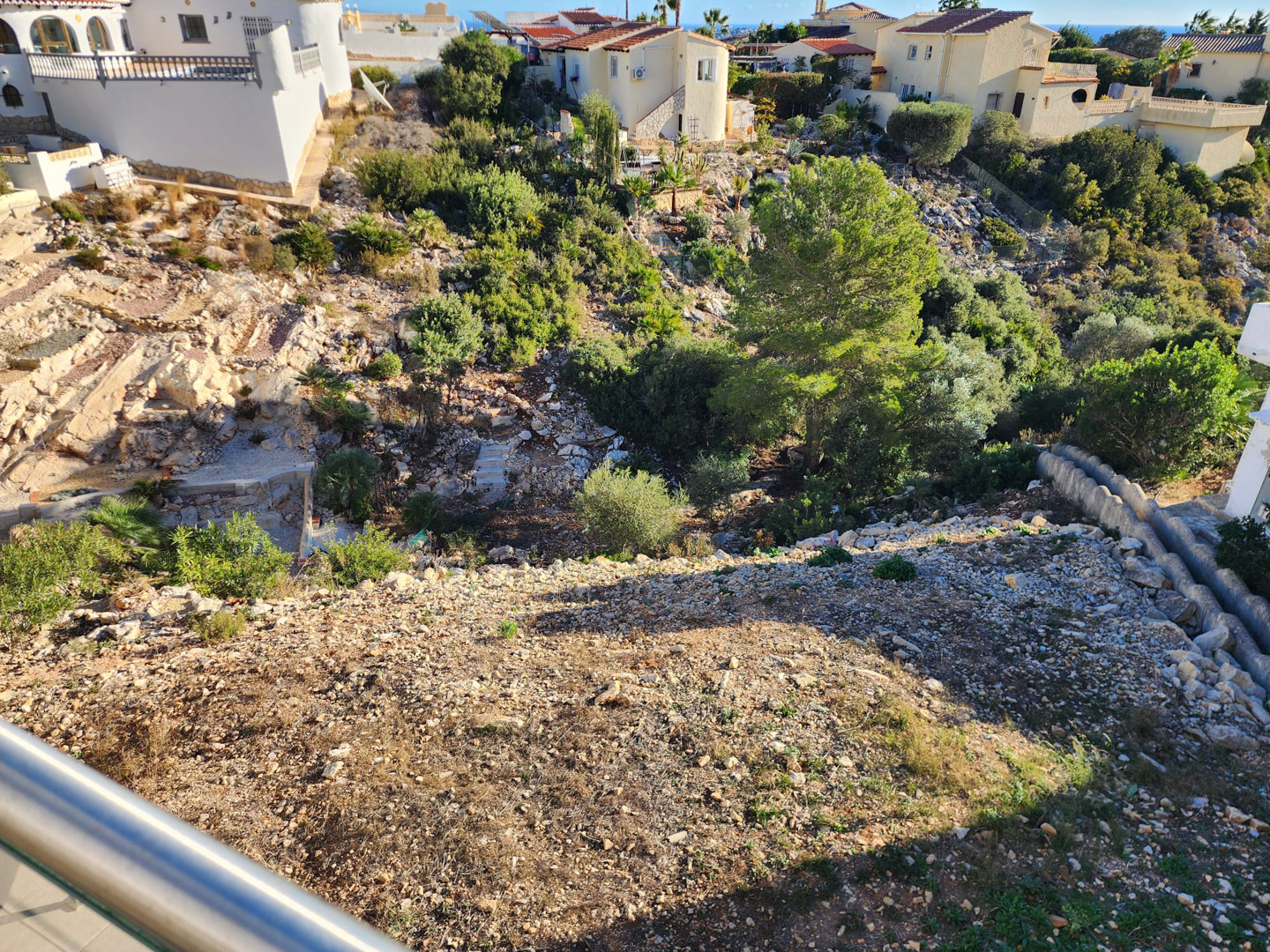 Uitzicht vanaf het balkon op het hellende perceel met grind, natuurlijke rotsformaties en inheemse mediterrane beplanting.