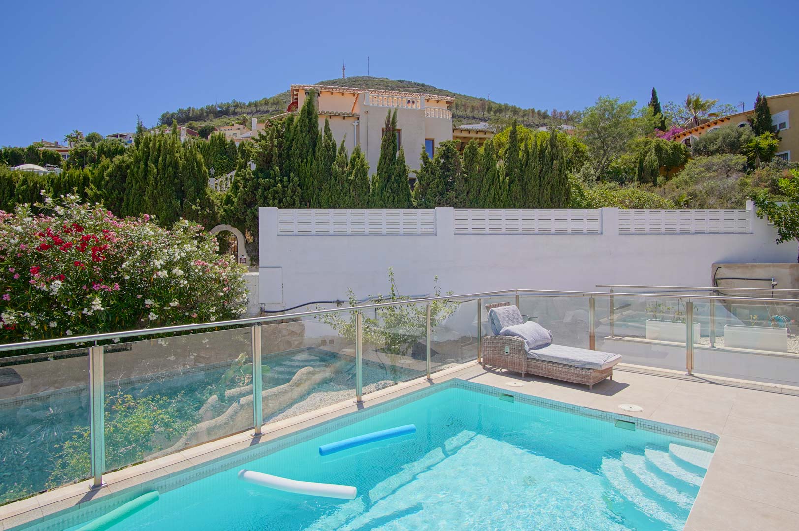 Zonnig terras met rechthoekig zwembad, glazen balustrades en een rotan ligbed, omgeven door mediterrane beplanting zoals oleanders en cipressen.
