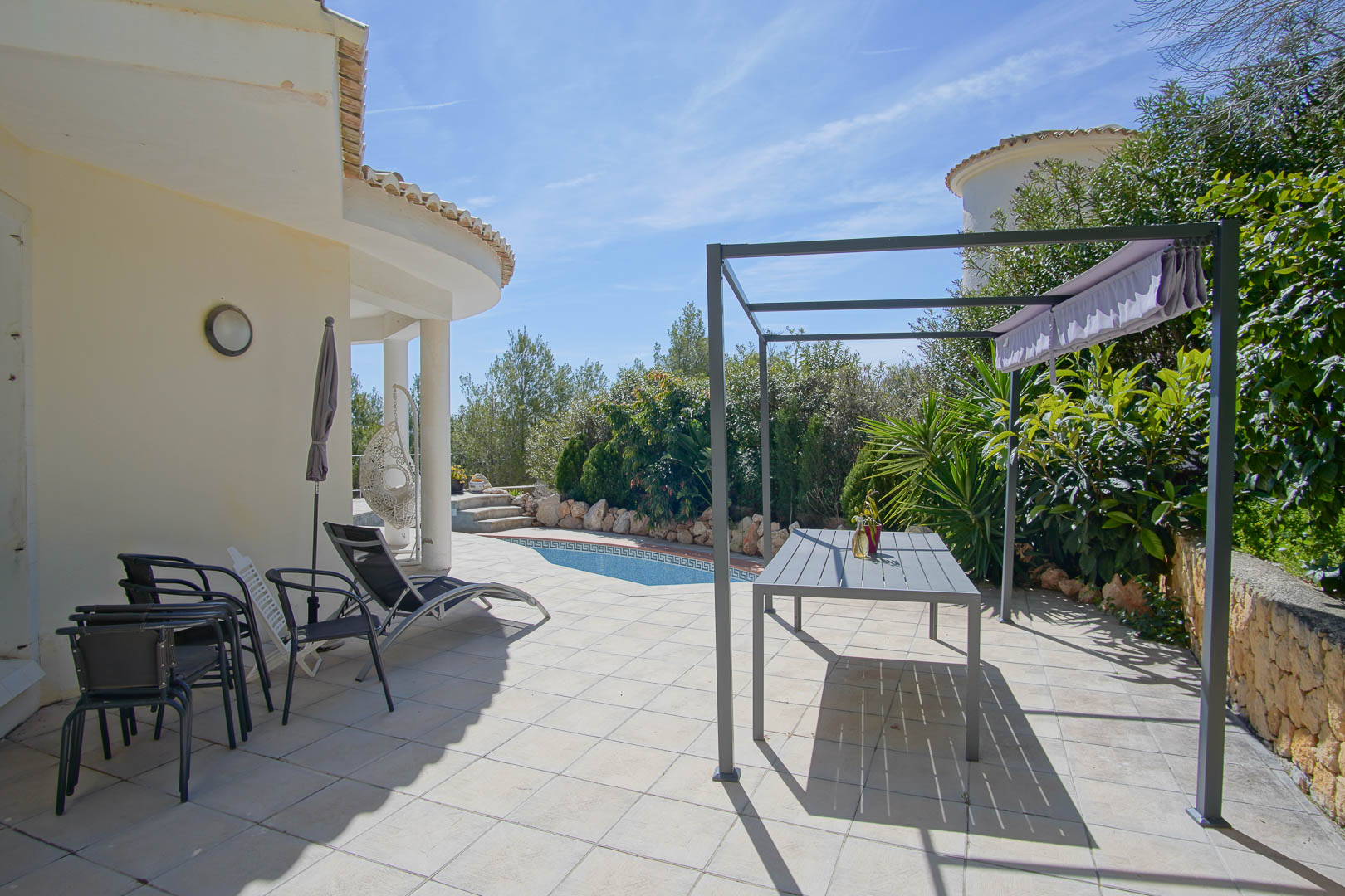 Betegeld buitenterras met een metalen pergola, eethoek en privézwembad omgeven door volwassen mediterrane beplanting.