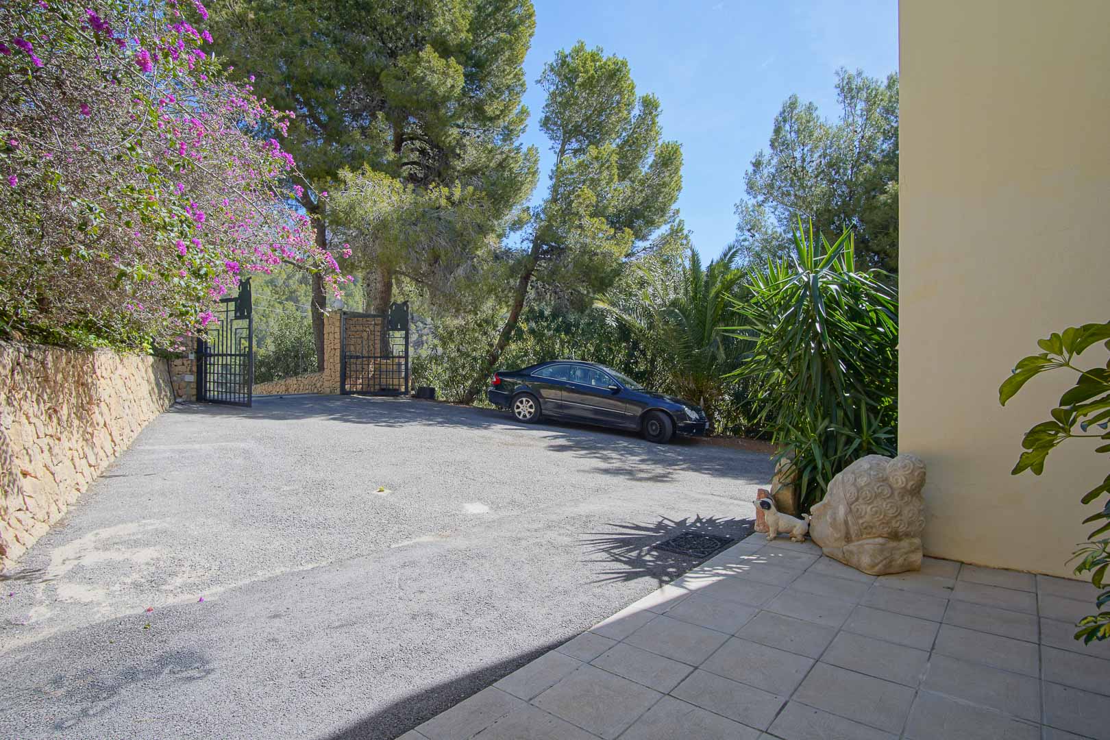 Beveiligde entree met ijzeren poort, ruime verharde parkeergelegenheid en natuurstenen muren omgeven door pijnbomen en mediterrane planten.
