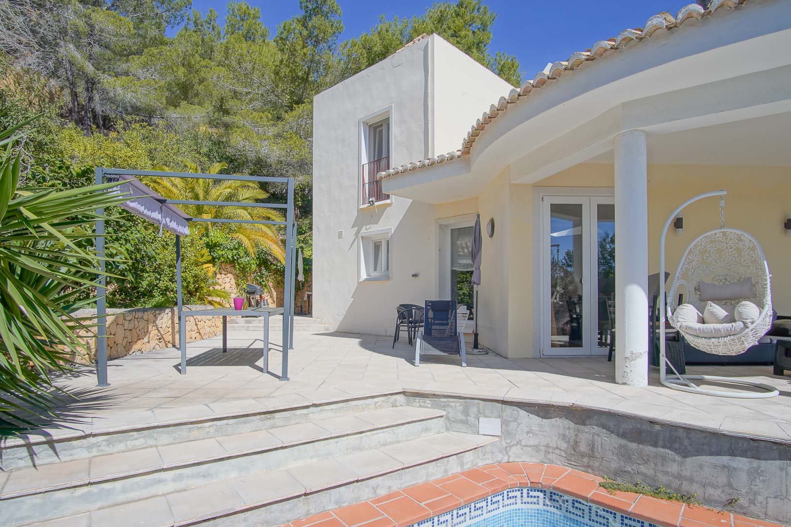 Buitenterras met zwembadrand, een eethoek onder de pergola en een overdekte veranda met hangstoel, omgeven door mediterrane begroeiing en pijnbomen.